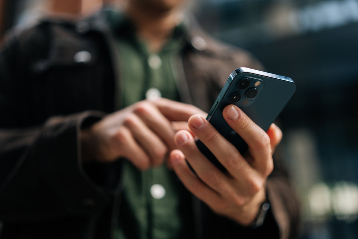 A person wearing a green shirt and brown jacket is using a smartphone outdoors. The focus is on the hands and phone, with the background blurred.