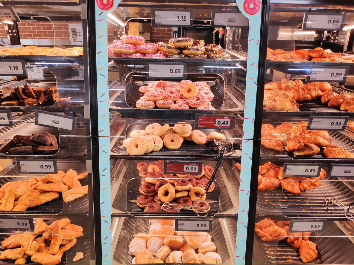 A bakery display case filled with assorted pastries, donuts, croissants, and baked goods arranged on trays, each with visible price tags. The pastries are organized in rows behind glass.