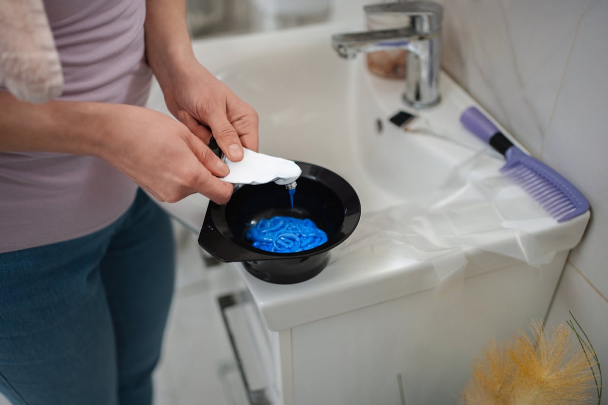 A person stands by a bathroom sink, squeezing blue hair dye from a tube into a black mixing bowl. Practicing hotel room etiquette, they use a white towel to protect the counter while a purple comb and hair coloring brush rest on the sink nearby.