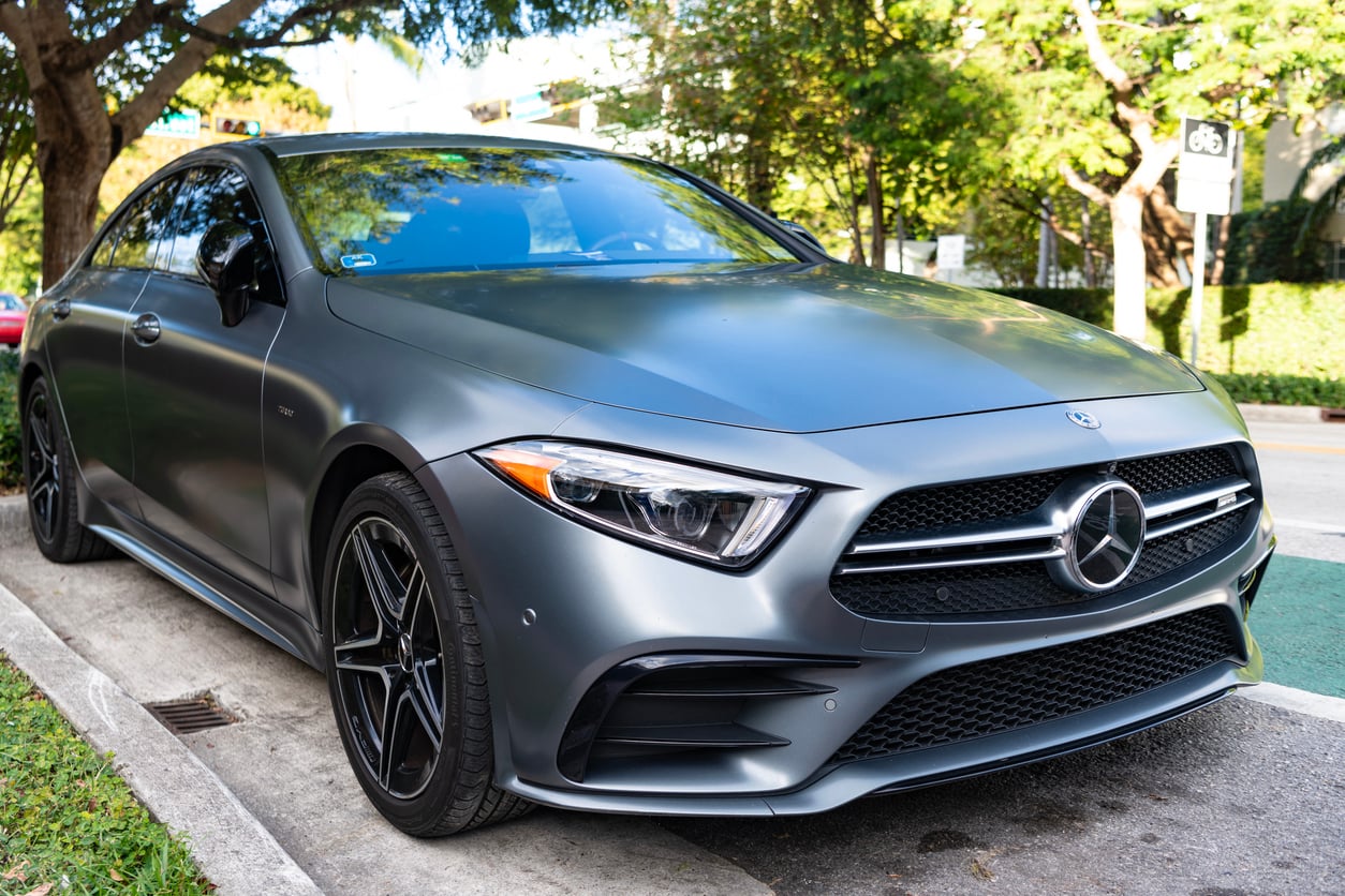 A matte gray Mercedes-Benz sedan is parked on the side of a street, surrounded by trees and greenery on a sunny day.