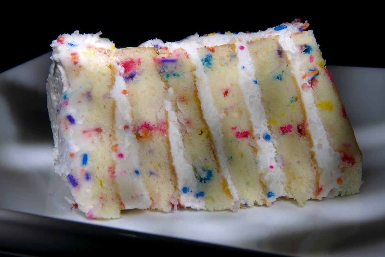 A close-up of a slice of funfetti cake with white frosting and colorful sprinkles, showing multiple layers on a white plate against a dark background.