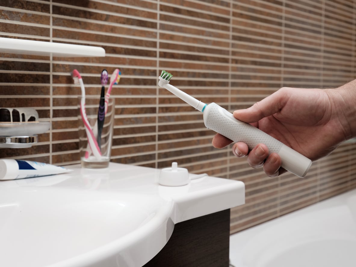 A hand holding a white electric toothbrush over a bathroom sink, with manual toothbrushes in a cup and toothpaste on the counter in the background. Brown striped tiles cover the bathroom wall.