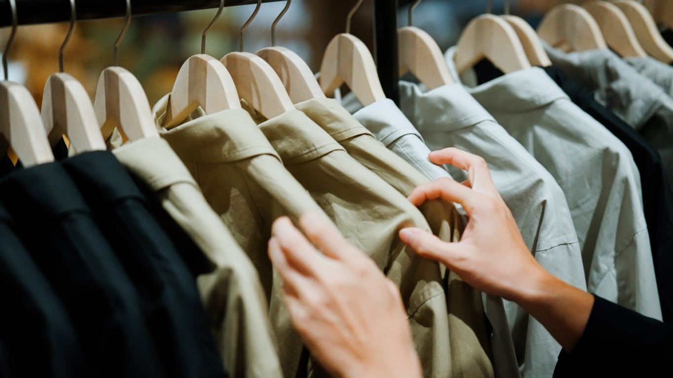 A person browses through a row of long-sleeve shirts on wooden hangers, selecting from a variety of neutral colors like black, beige, and white.