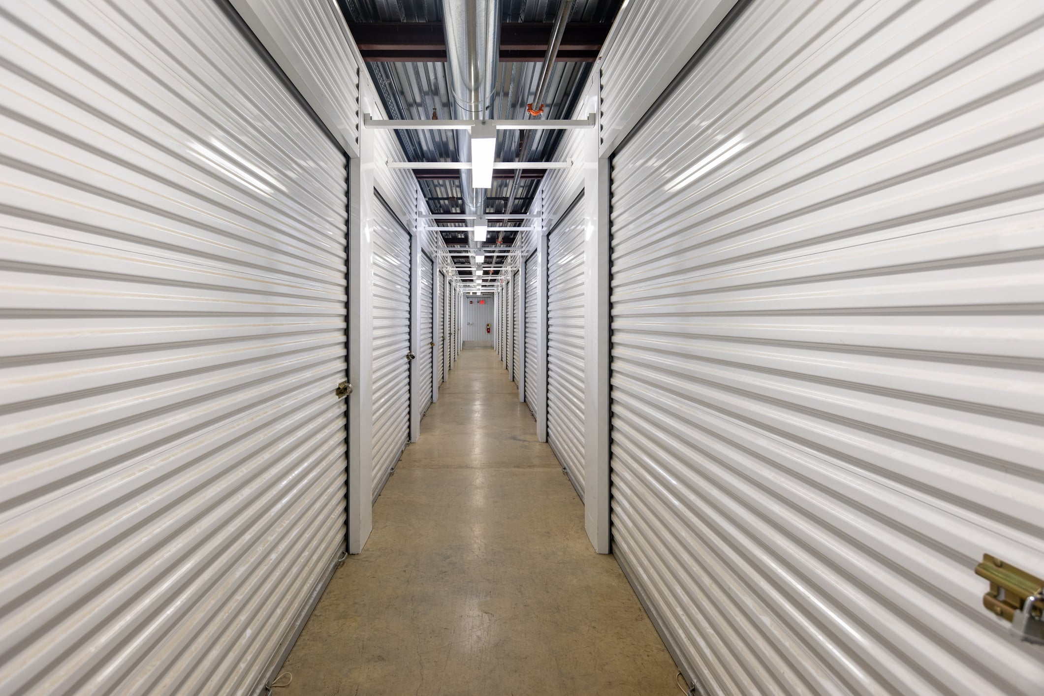 A brightly lit hallway lined with white metal storage unit doors on both sides, featuring a concrete floor and overhead pipes, in a self-storage facility.
