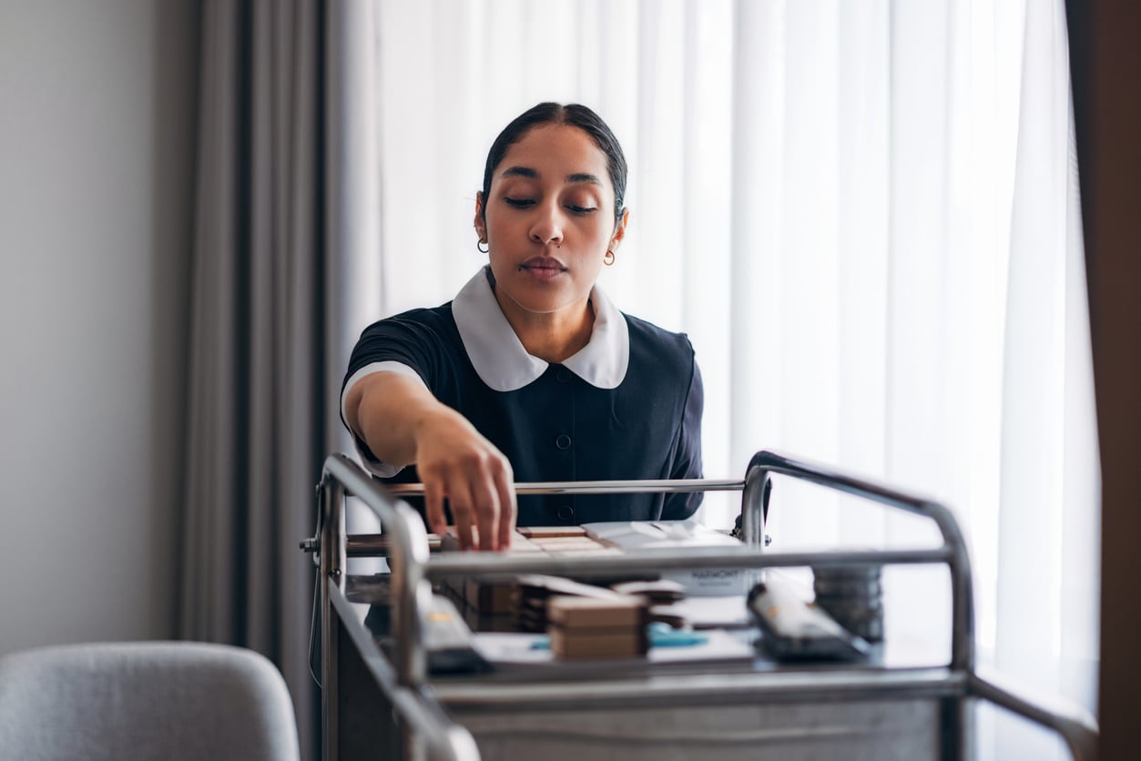 A woman in a housekeeping uniform, focused on her task, arranges items on a cleaning cart in a bright room with large windows and sheer curtains, demonstrating proper hotel room etiquette.