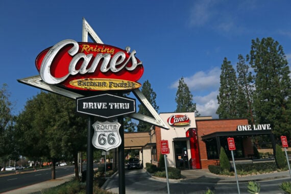 A Raising Cane&rsquo;s Chicken Fingers restaurant with a large sign in front reading &ldquo;Drive Thru&rdquo; and &ldquo;Route 66.&rdquo; The restaurant building and several trees are visible under a blue sky.