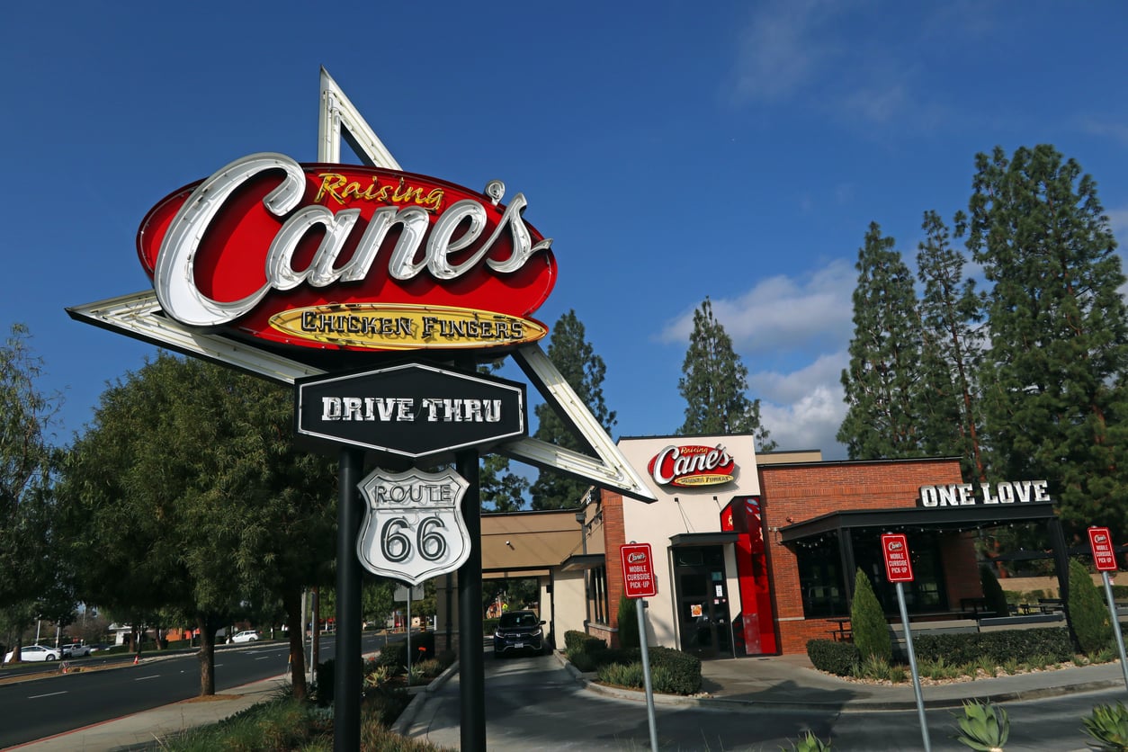 A Raising Cane’s Chicken Fingers restaurant with a large sign in front reading “Drive Thru” and “Route 66.” The restaurant building and several trees are visible under a blue sky.