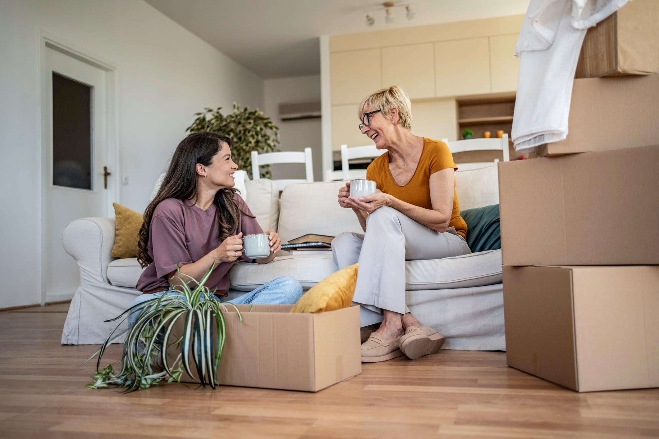 Two women smiling and talking while holding mugs, surrounded by moving boxes in a living room, discuss how to support adult children financially. One woman sits on the couch, the other on the floor next to an open box with a potted plant.