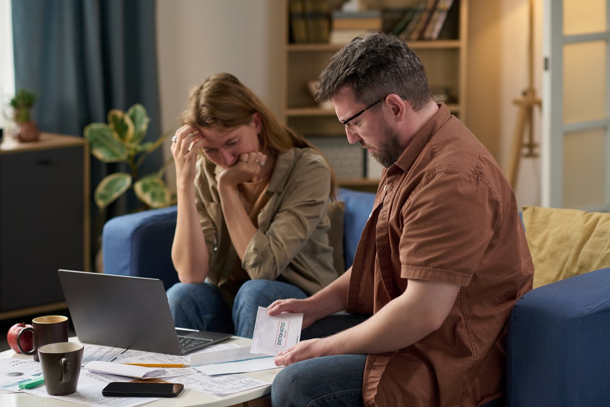 A man and woman sit on a couch looking stressed while reviewing bills. The man holds an envelope labeled "Overdue," and the woman rests her head on her hand. A laptop, phone, and documents are spread out on the table.