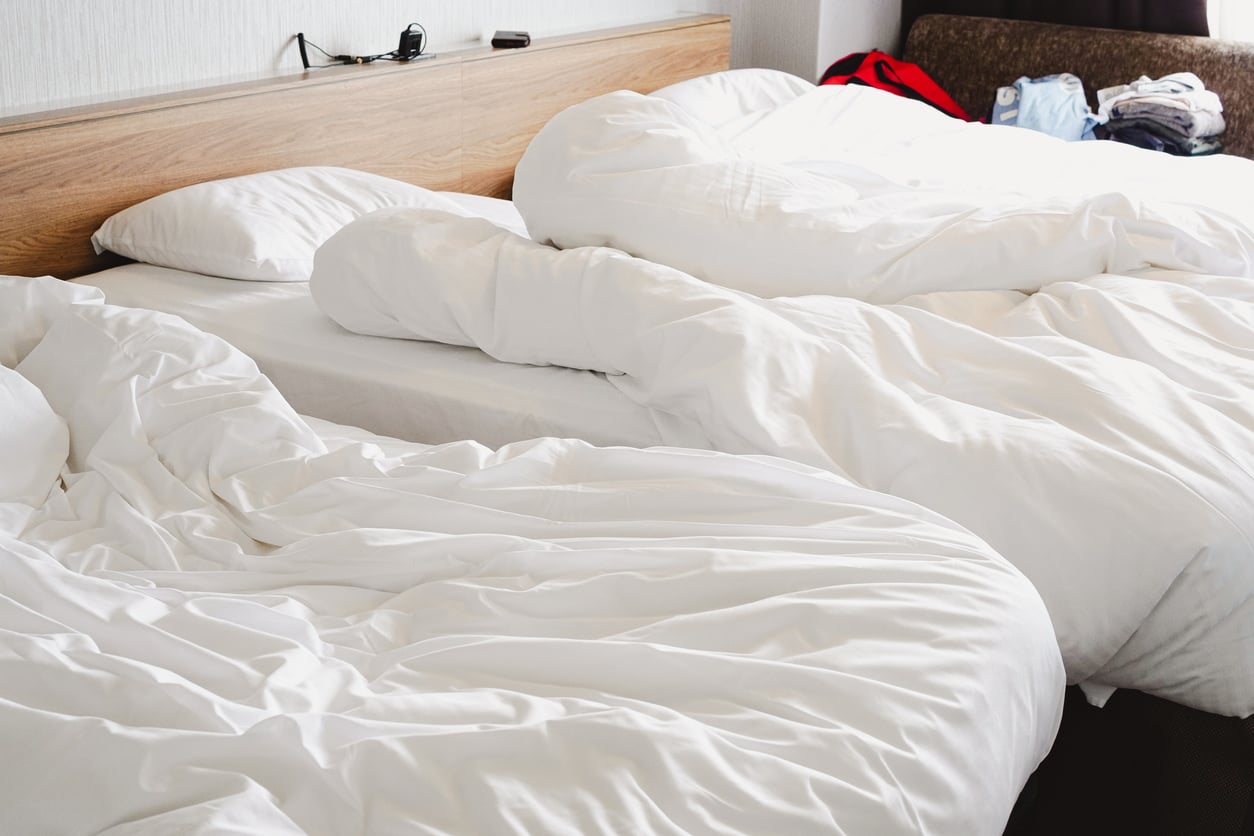 Two unmade beds with white sheets and pillows, set against a wooden headboard. Clothes are visible in the background on a brown sofa—a gentle reminder of hotel room etiquette. A charger with cables rests on the headboard shelf.