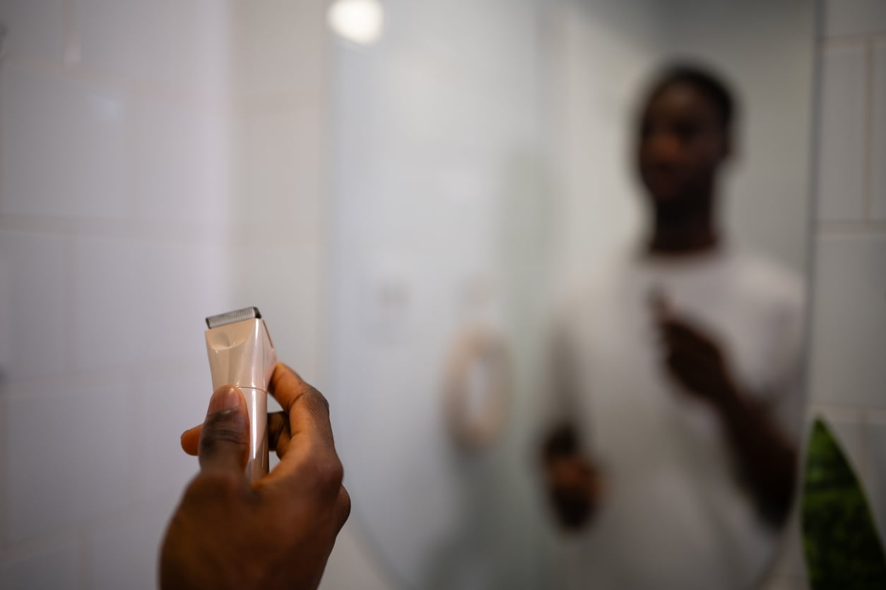 A person holds electric hair clippers in front of a bathroom mirror, their blurred reflection visible in the background, suggesting preparation for grooming or shaving while mindful of hotel room etiquette.