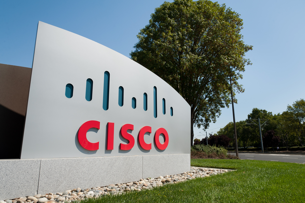 A large outdoor Cisco sign with red lettering and a blue logo stands on a landscaped area near a road, set against news of potential 2026 layoffs, with a tree and clear blue sky in the background.
