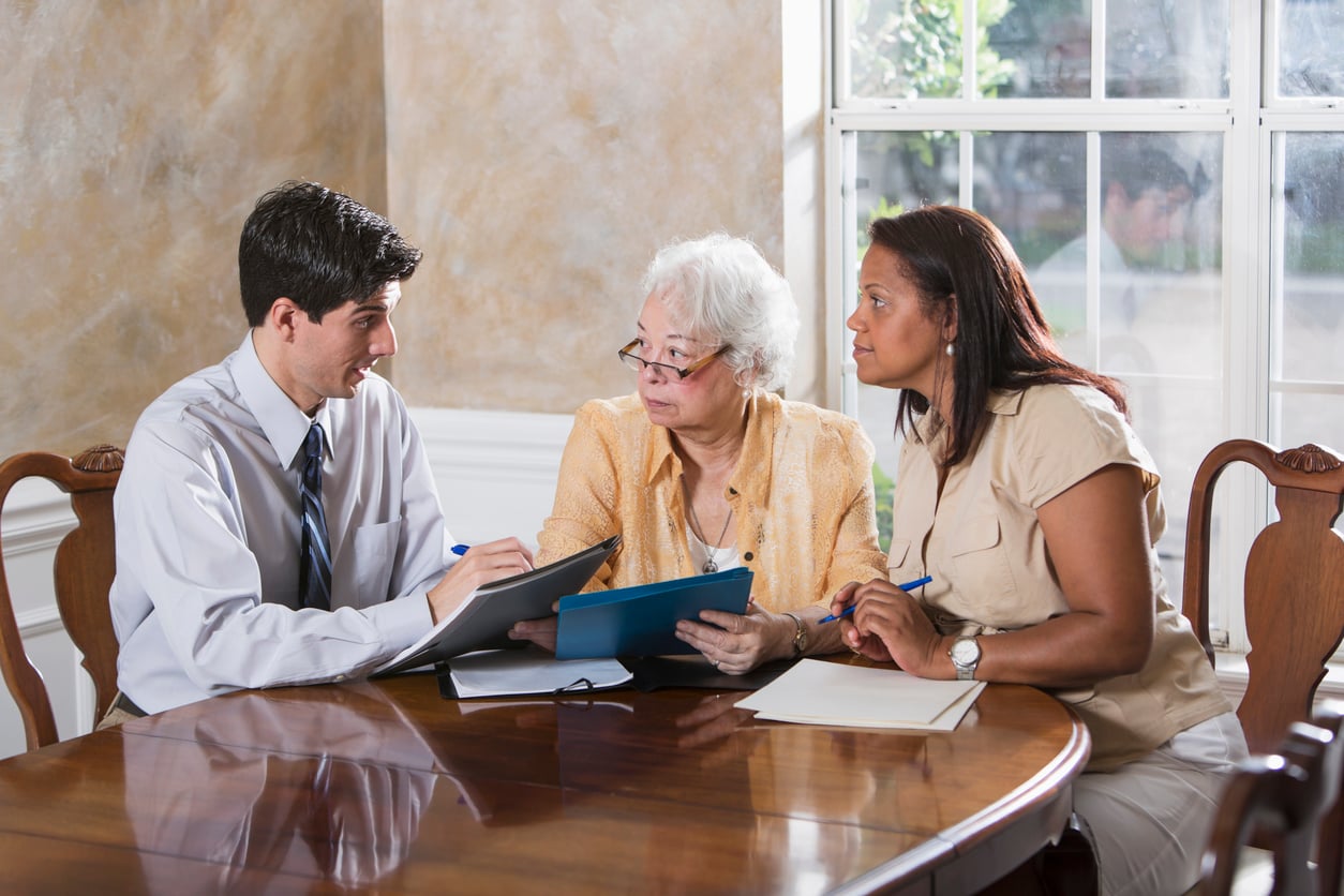 A man in a suit shows documents to two women sitting at a table. One woman is elderly and holding a clipboard, while the other is younger, listening attentively. Sunlight streams in through a large window behind them.