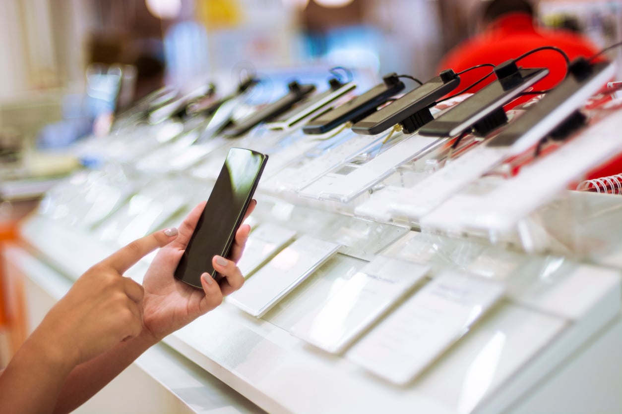 A person’s hand tests a smartphone on display at an electronics store, with several phones lined up on a counter, each secured with security devices.
