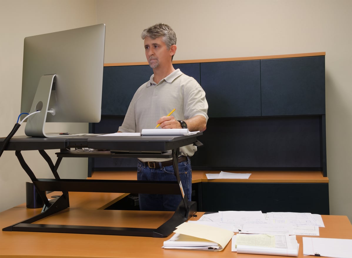 A man stands at a desk using a computer with a standing desk converter. He is writing in a notebook, surrounded by papers and folders in a modern office setting.