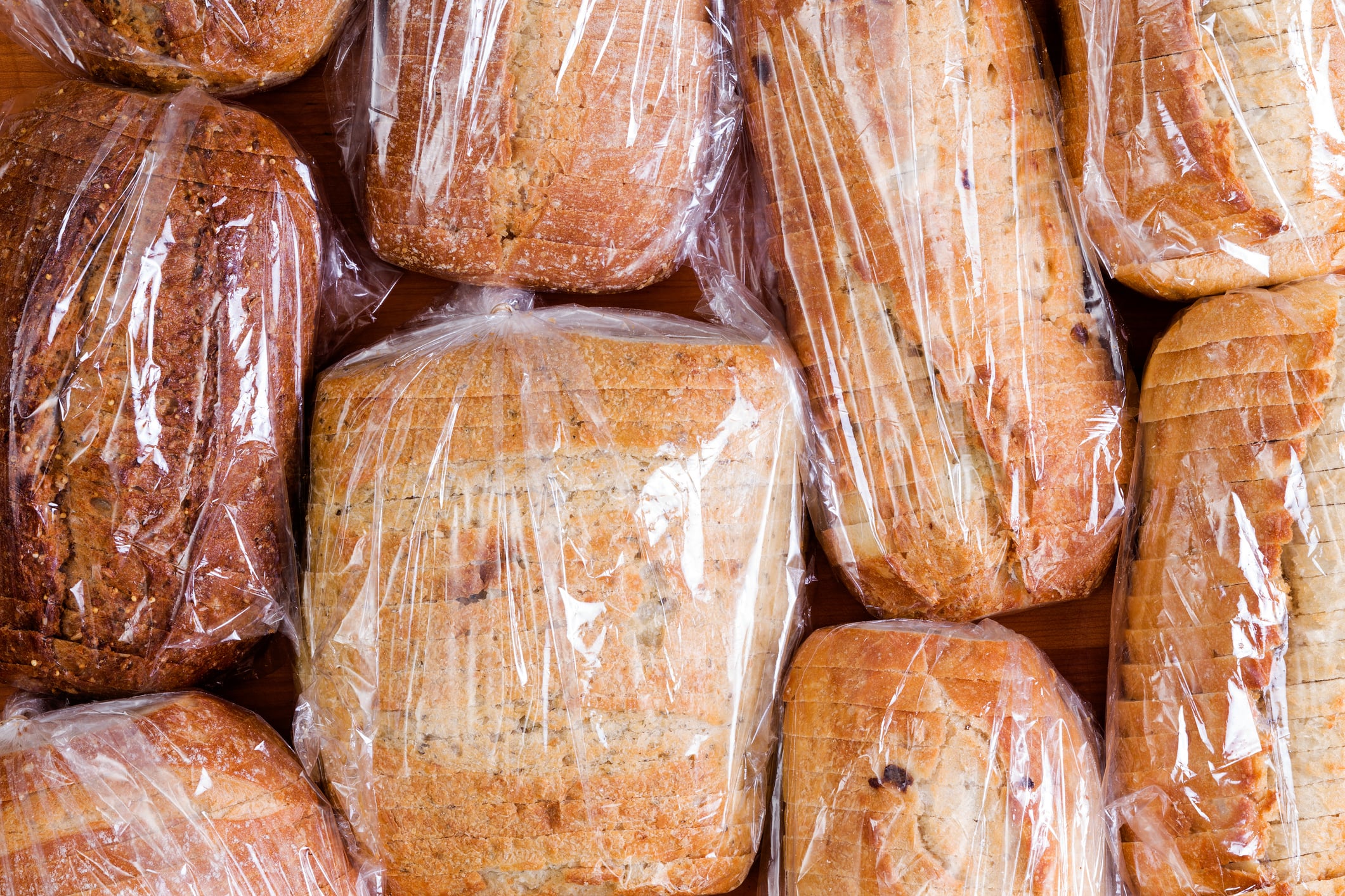 Several loaves of sliced bread are wrapped in clear plastic and arranged closely together on a wooden surface, showing different varieties and crust colors.