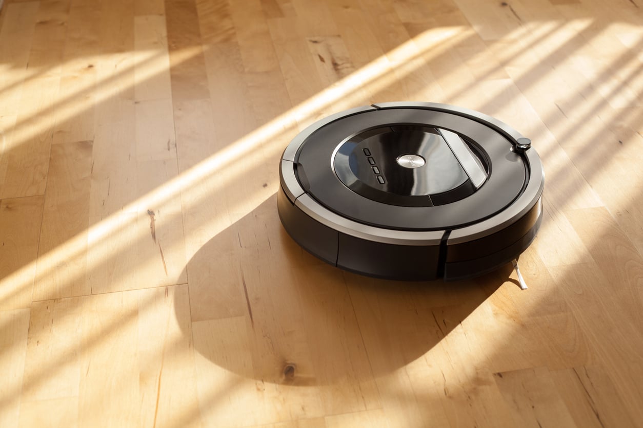 A black robotic vacuum cleaner moves across a wooden floor in sunlight, casting a shadow along the light-colored planks.