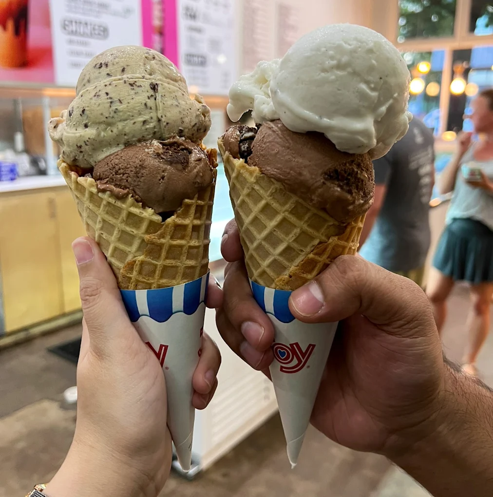 Two hands holding waffle cones with two scoops of ice cream each; one cone has chocolate and cookies & cream, the other has chocolate and vanilla. Blurred ice cream shop background with people standing inside.