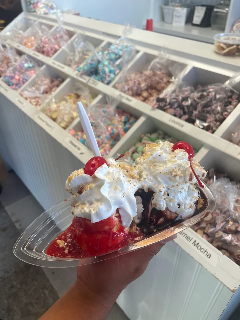 A hand holds a banana split sundae topped with whipped cream and cherries in front of a candy store display, capturing the sweet magic you'd find at the best ice cream shop in every state.