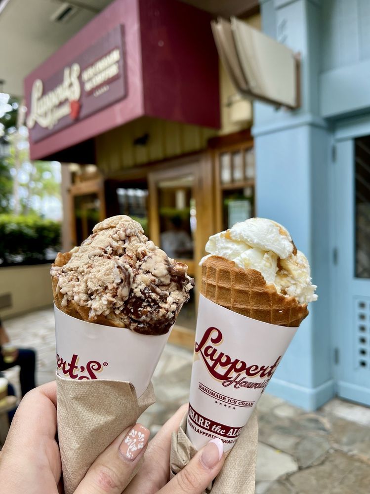 Two hands hold waffle cones with creamy scoops in front of Lappert's, known as the best ice cream shop in every state. The left cone has a chocolate and nut flavor; the right features caramel or vanilla. Both cones have branded Lappert’s wrappers.