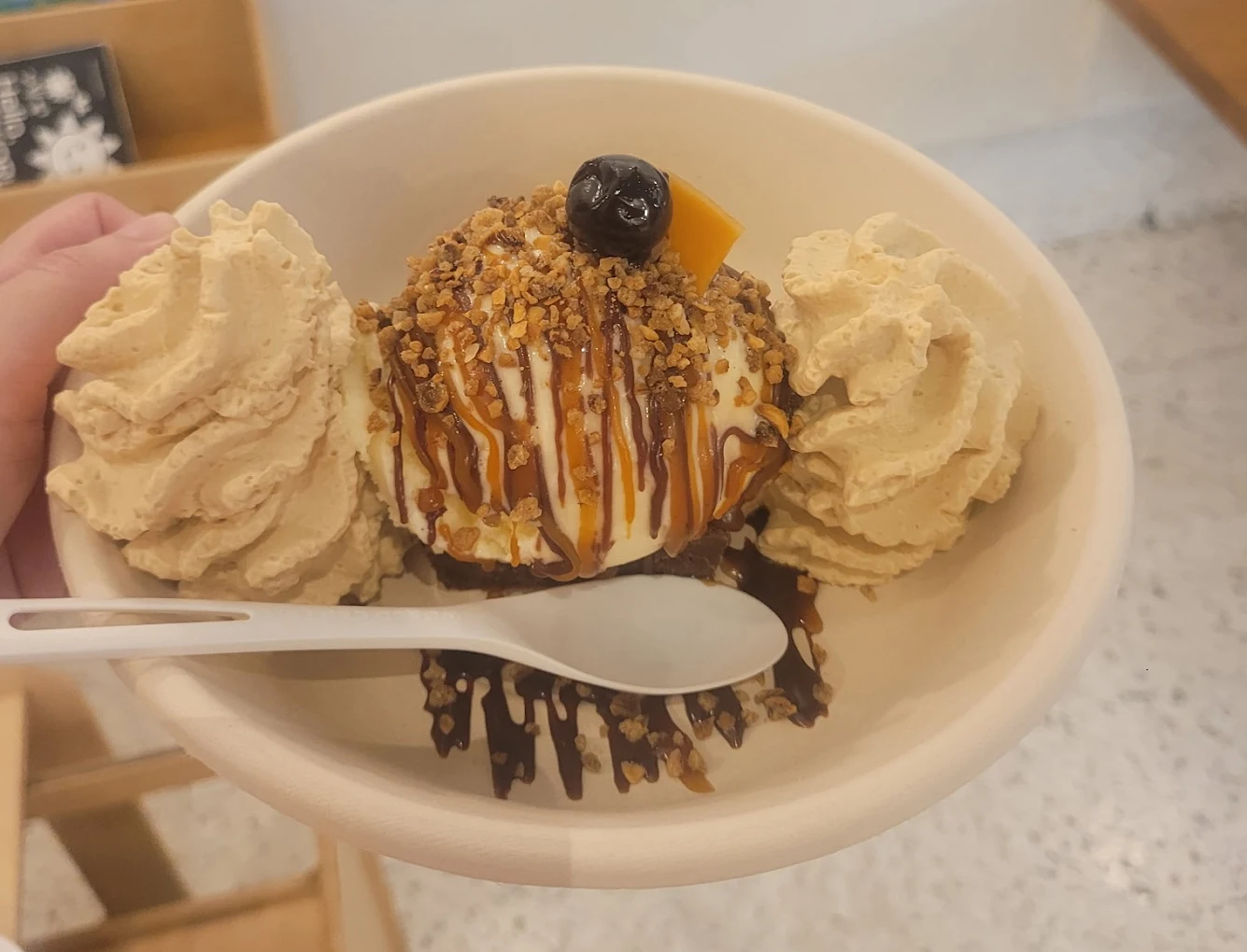 A bowl of ice cream topped with caramel sauce, crushed nuts, a cherry, and a small orange piece, flanked by two swirls of whipped cream, with a white spoon resting on the bowl.