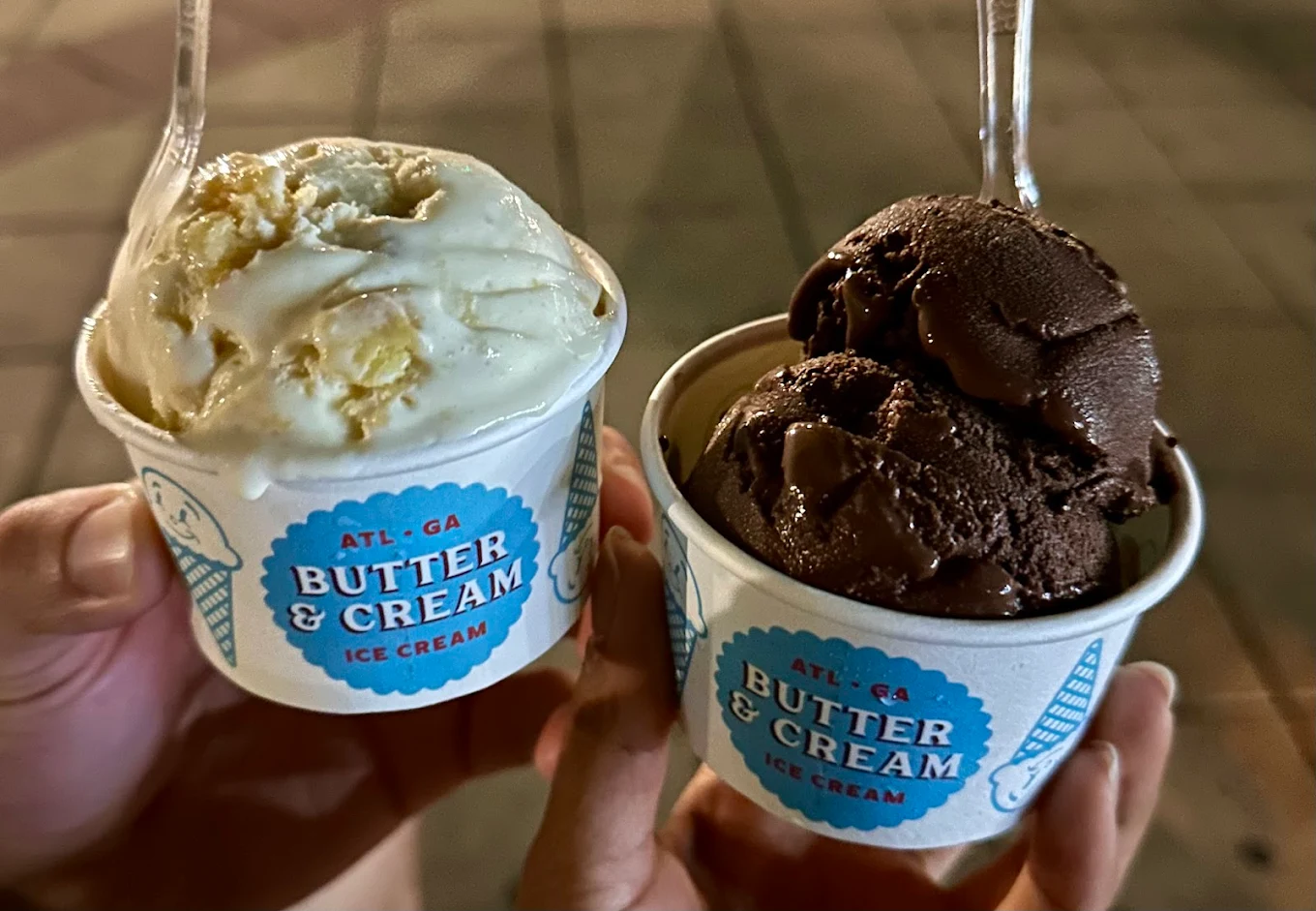 Two hands holding cups of Butter & Cream ice cream; one cup has two scoops of vanilla and the other has two scoops of chocolate. Both cups have clear plastic spoons. The background appears to be a sidewalk.