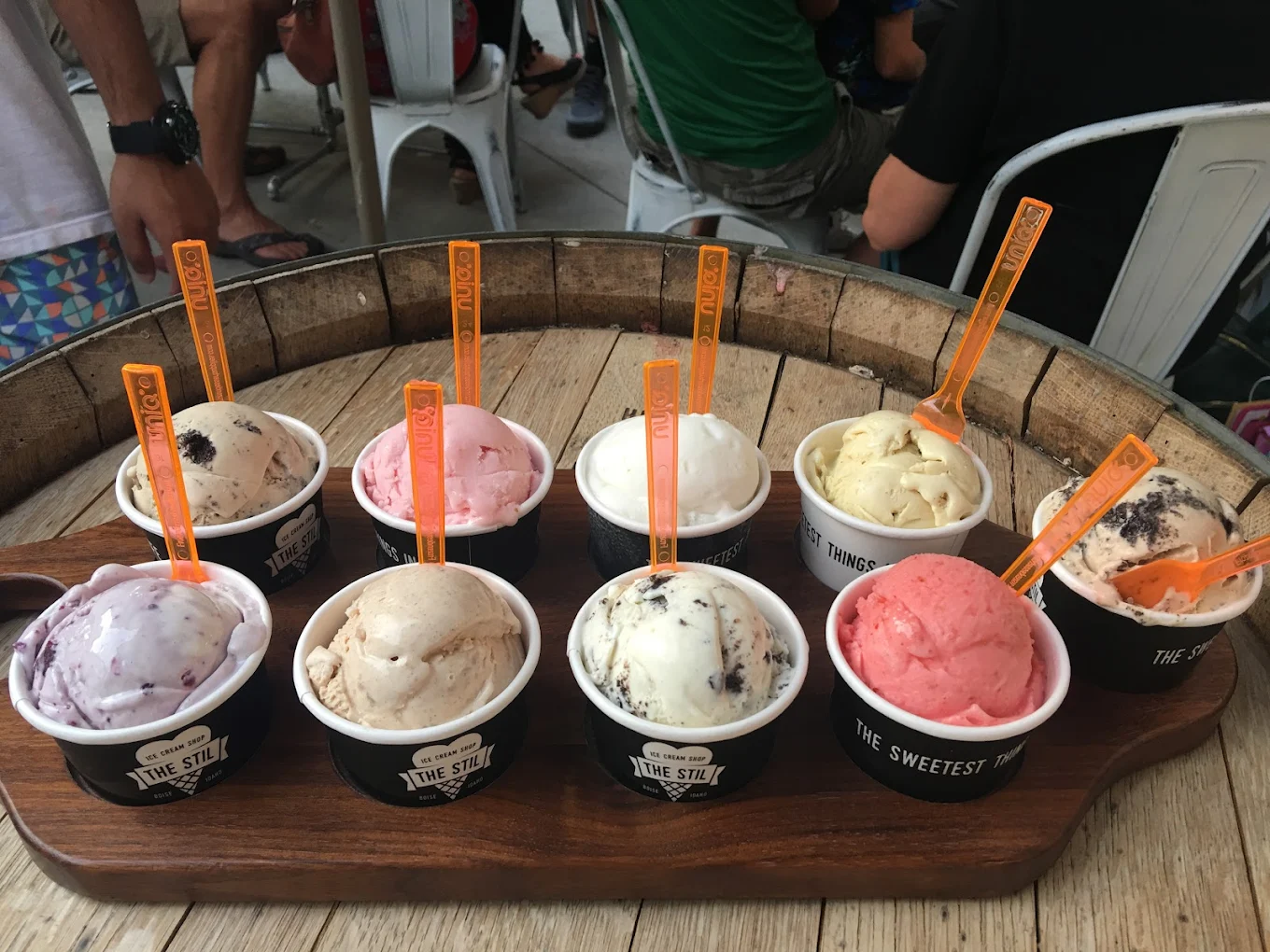 Nine cups of assorted ice cream flavors are arranged on a wooden tray, each with an orange spoon, set on a rustic wooden table with people seated in the background.