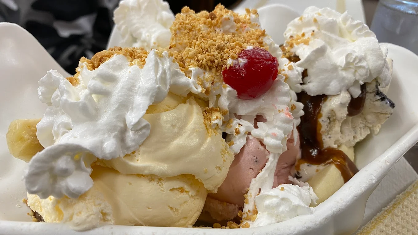 A close-up of a banana split topped with vanilla, strawberry, and chocolate ice cream, whipped cream, crushed nuts, a maraschino cherry, and chocolate syrup in a white dish.