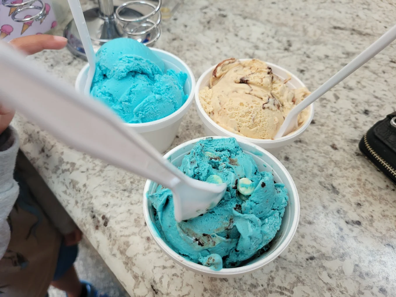 Three cups of ice cream on a marble countertop: one with bright blue ice cream, one with light brown ice cream with chocolate swirls, and another with blue ice cream with chocolate pieces. Each cup has a white plastic spoon.