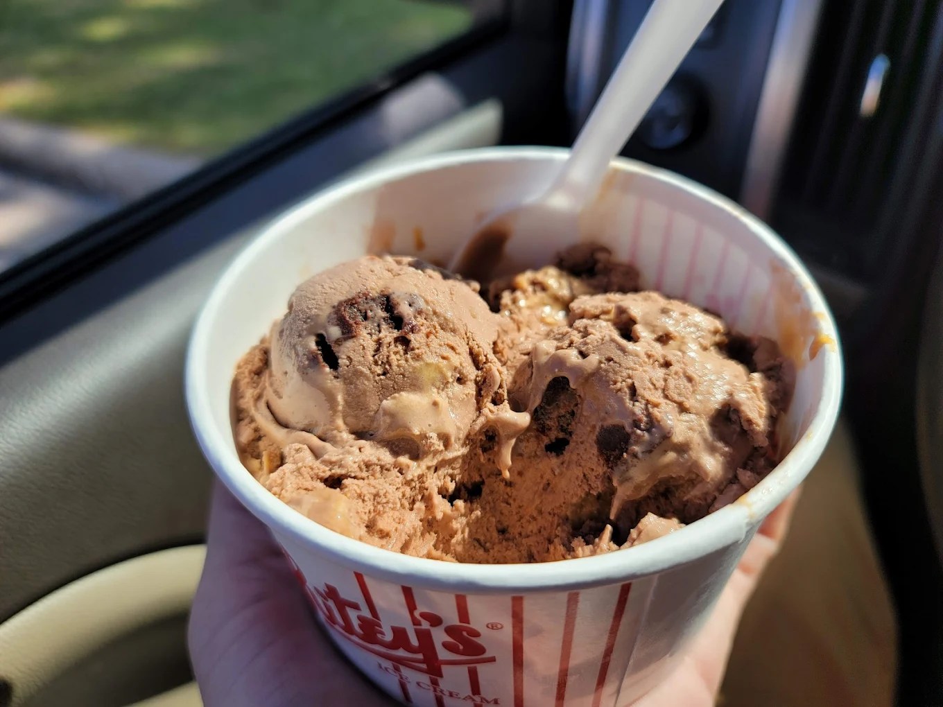 A hand holds a cup filled with scoops of chocolate ice cream with chunks, and a white plastic spoon, inside a car with sunlight shining through the window.