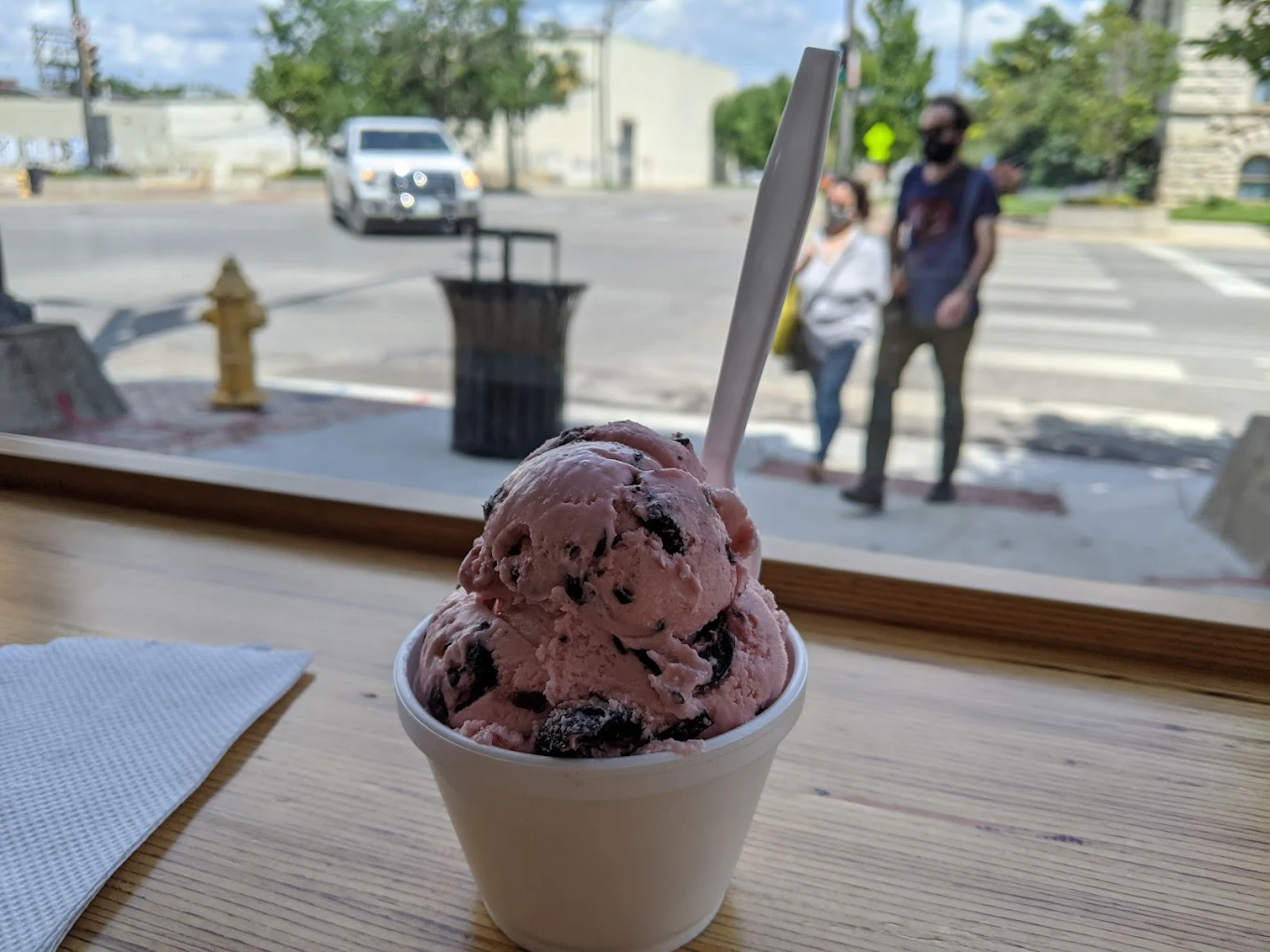 A cup of pink ice cream with dark chunks and a white spoon sits on a wooden table by a window, with a napkin nearby. Outside, a street and two blurred people walking are visible in the background.