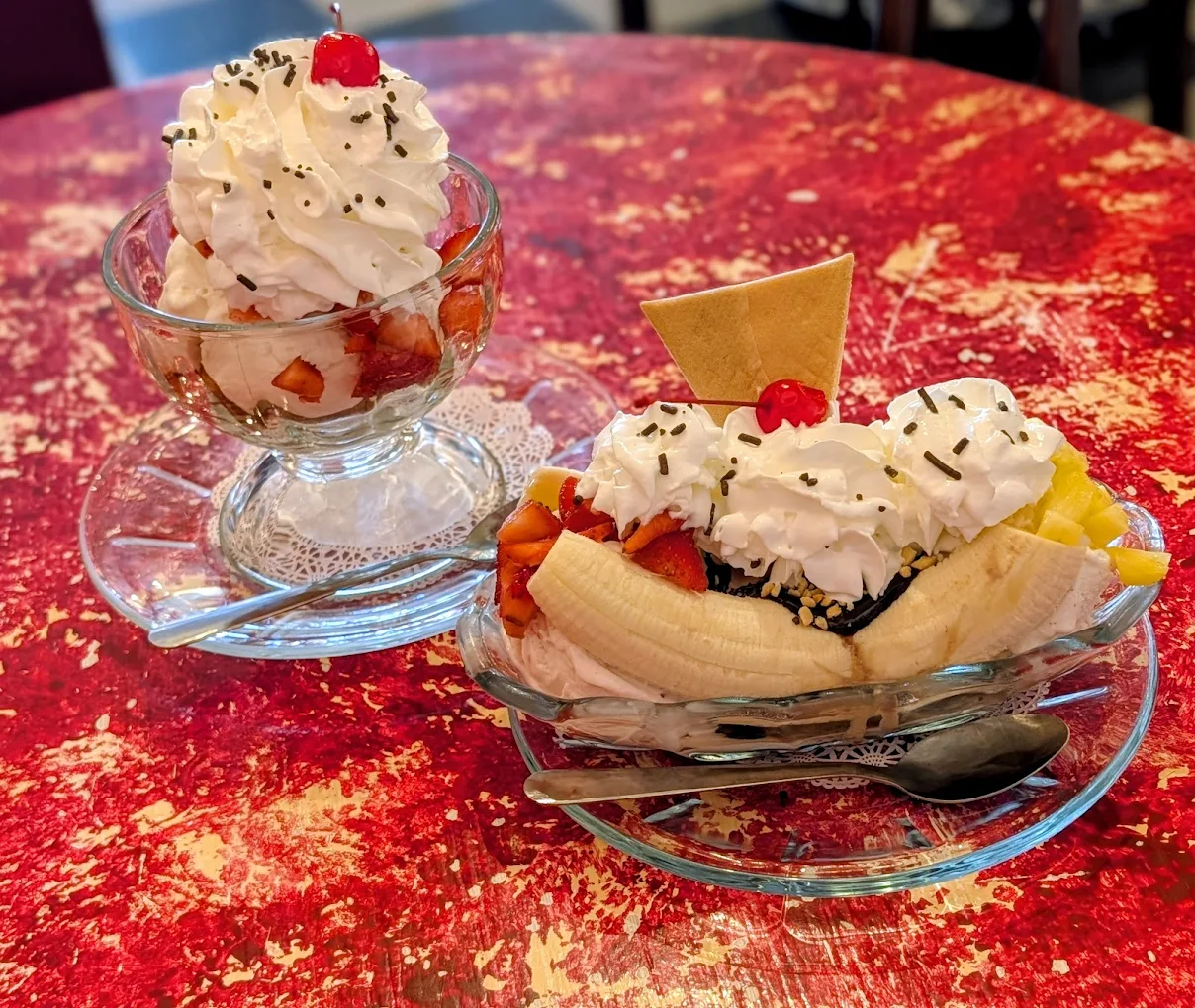 Two glass dishes on a red textured table: one holds a banana split with whipped cream, sprinkles, fruit, and a cherry; the other has a sundae with cream, sprinkles, fruit, and a cherry on top.