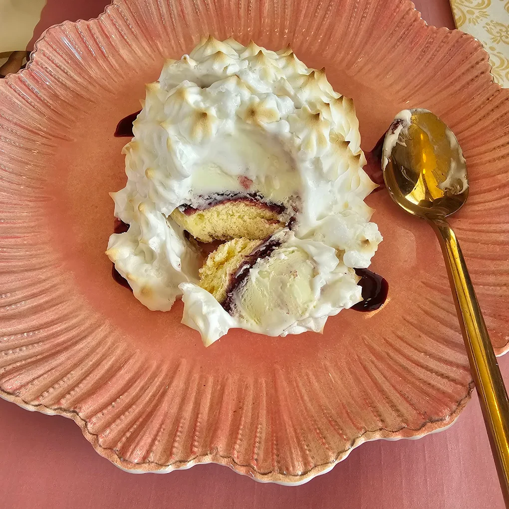 A partially eaten baked Alaska dessert with layers of cake, ice cream, and meringue sits on a textured pink plate beside a gold spoon.