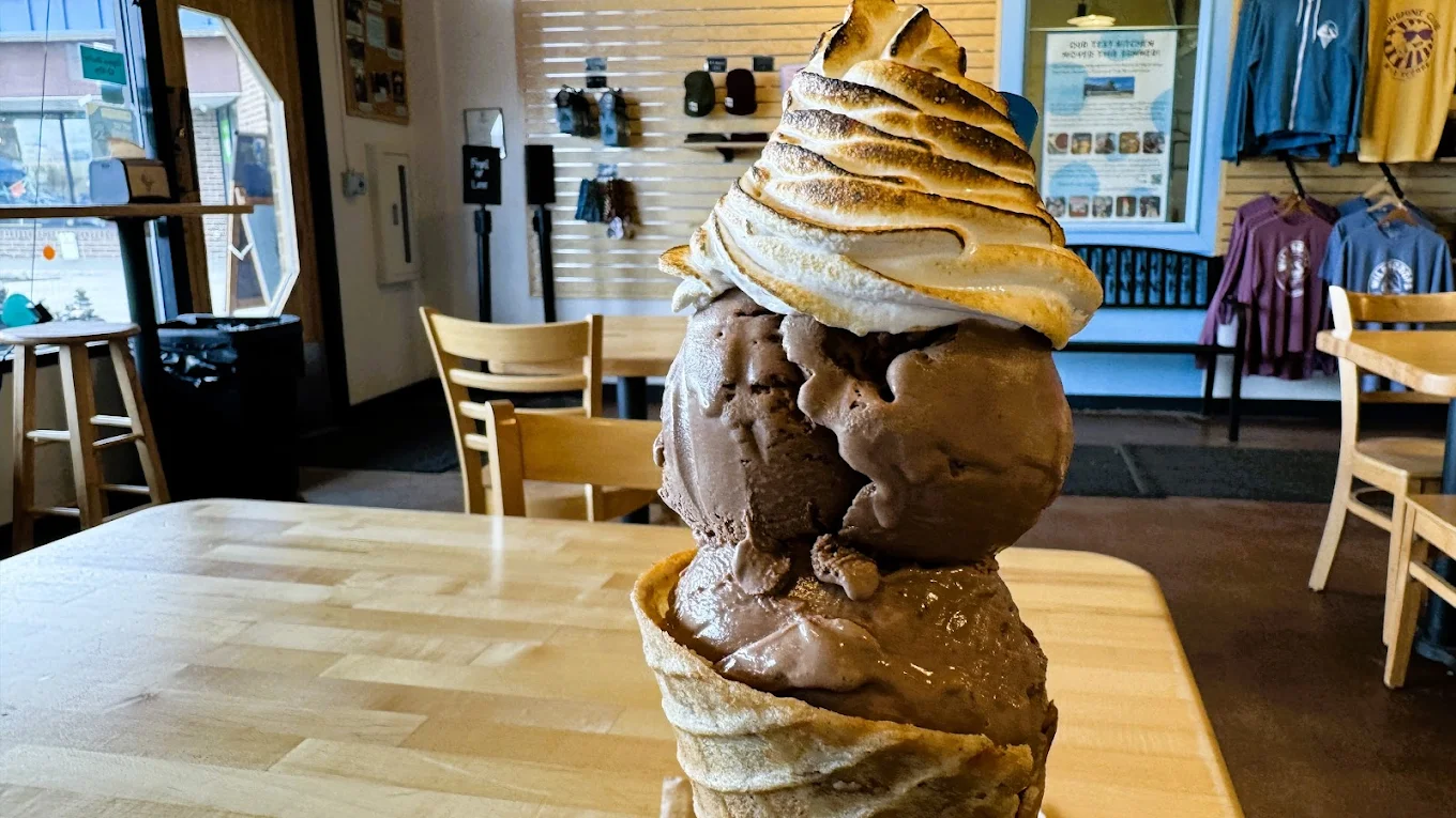 A large ice cream cone with multiple scoops of chocolate ice cream and a swirl of toasted meringue on top sits on a wooden table inside a cozy, casual shop with wooden chairs and wall displays.