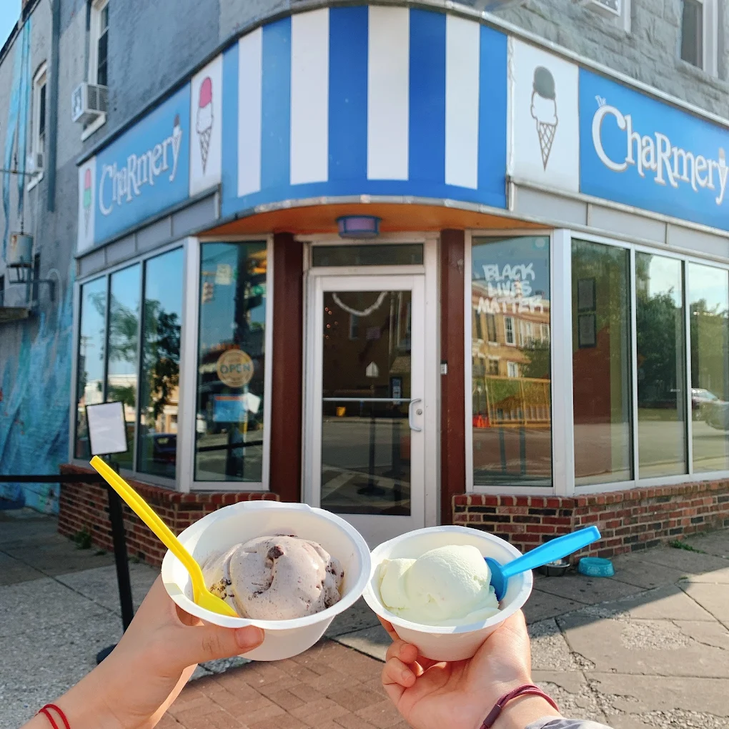 Two hands hold cups of ice cream with colorful spoons in front of The Charmery ice cream shop, which has a striped blue-and-white sign and a "Black Lives Matter" sign in the window.
