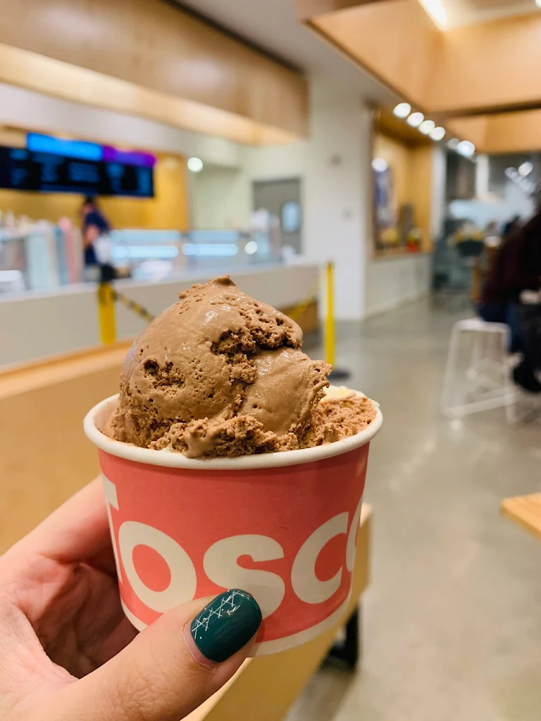 A hand with green painted nails holds a cup of chocolate ice cream inside a modern, brightly lit ice cream shop with wooden accents and blurred people in the background.