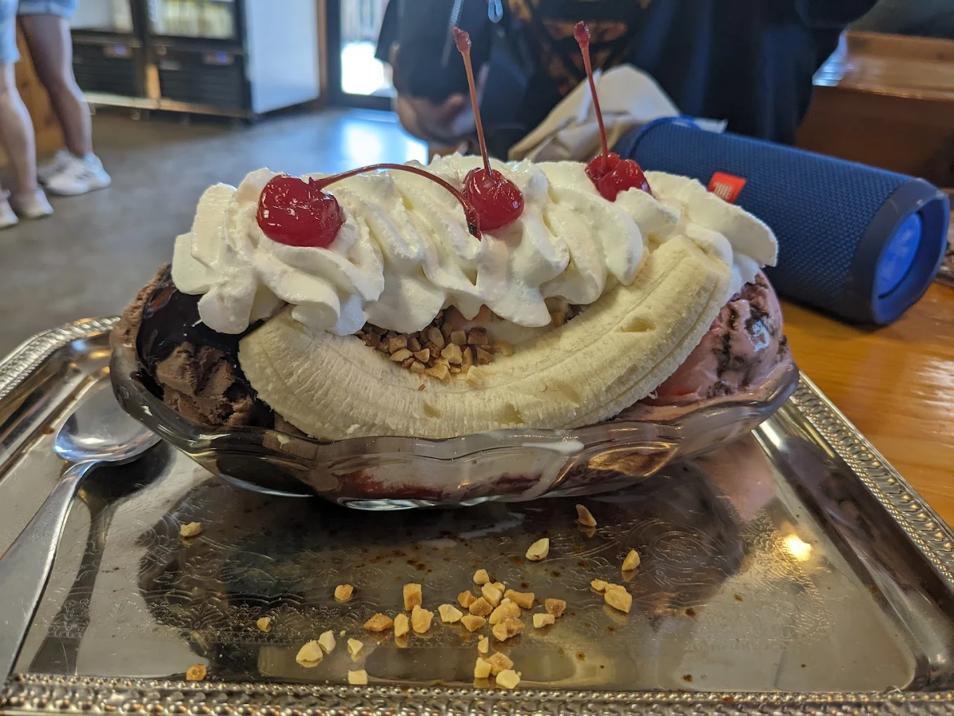 A banana split dessert with three scoops of ice cream, whipped cream, chopped nuts, and three cherries served in a glass dish on a silver tray. A spoon and a blue speaker are nearby.
