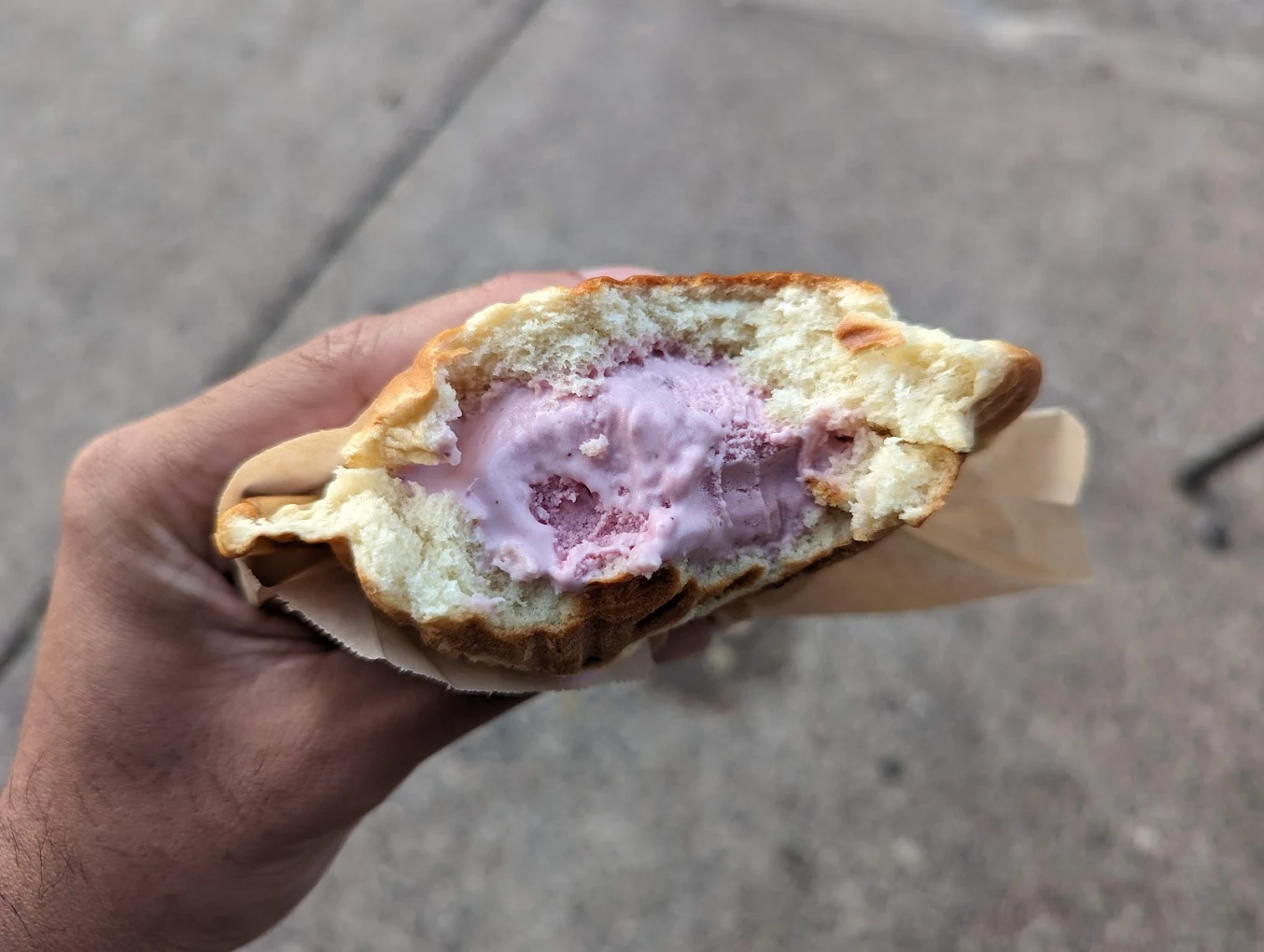 A hand holding a partially-eaten sandwich with pink ice cream filling, wrapped in brown paper, with a concrete sidewalk in the background.