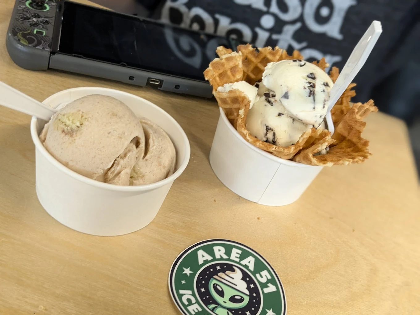Two servings of ice cream sit on a wooden table—one in a white cup, the other in a waffle bowl. A sticker with "AREA51 ICE" and a green alien logo is in front, with a gaming device and shirt partially visible in the background.