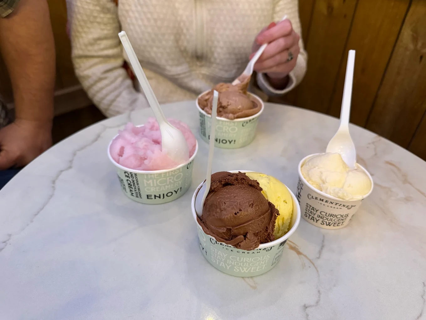 Four cups of ice cream in different flavors sit on a round white table, with two people—one in a cream sweater—reaching for their cups with plastic spoons. The setting appears to be indoors with wooden walls.