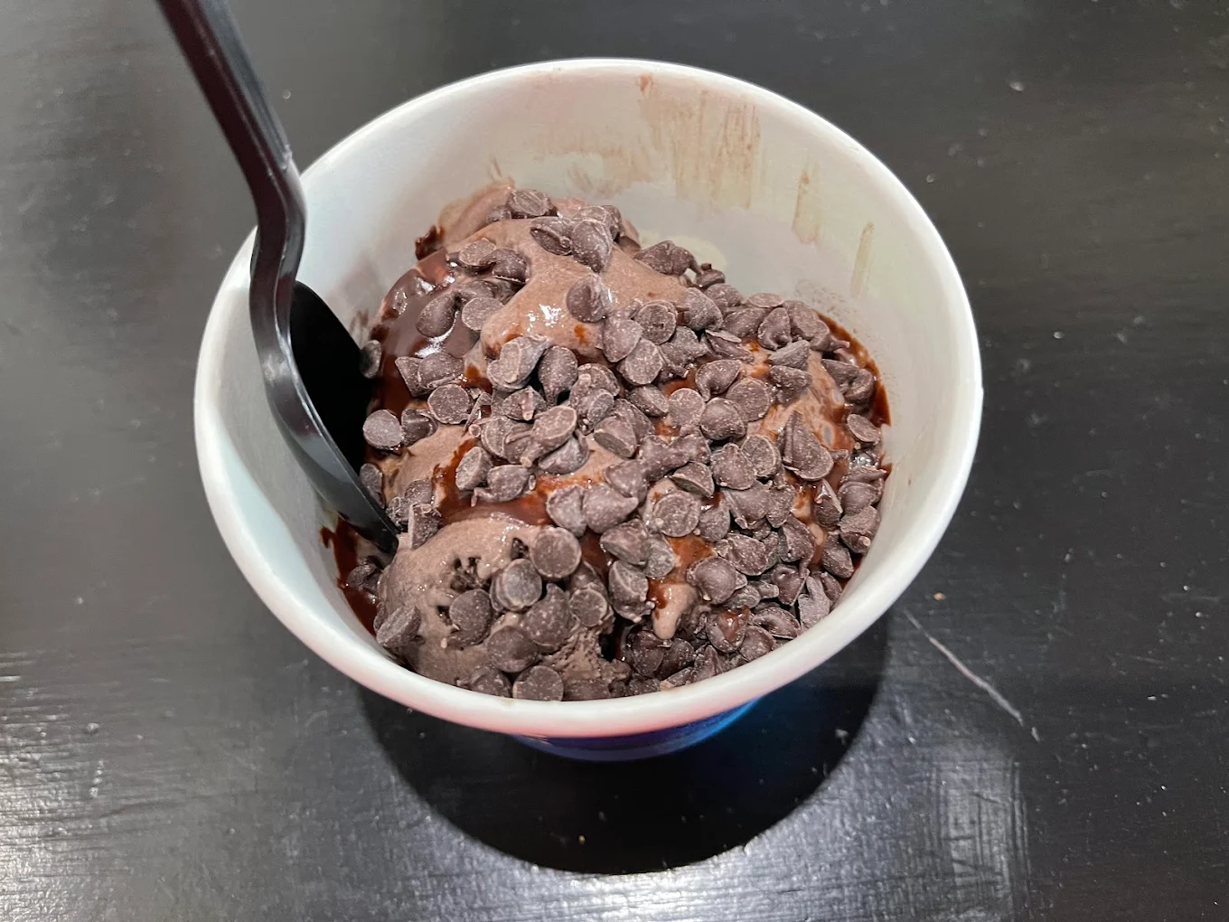 A cup of chocolate ice cream topped with chocolate chips and chocolate syrup, with a black plastic spoon, sits on a dark, glossy table.
