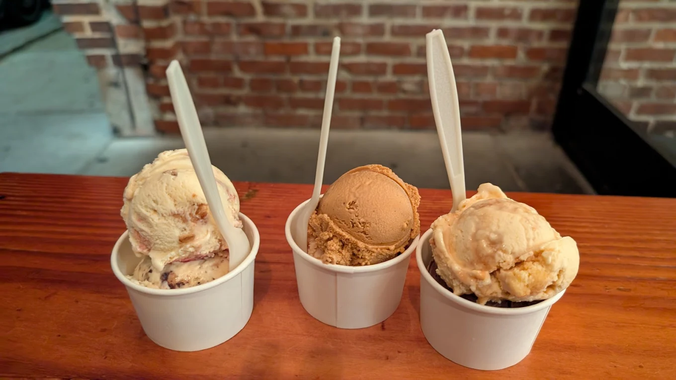 Three cups of ice cream with different flavors are placed side by side on a wooden table, each with a white plastic spoon. A brick wall is in the background.