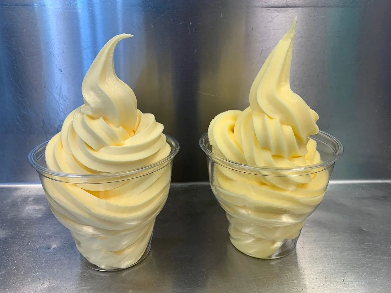 Two clear plastic cups filled with swirled yellow soft serve ice cream are placed side by side on a metallic surface, against a reflective metallic background.