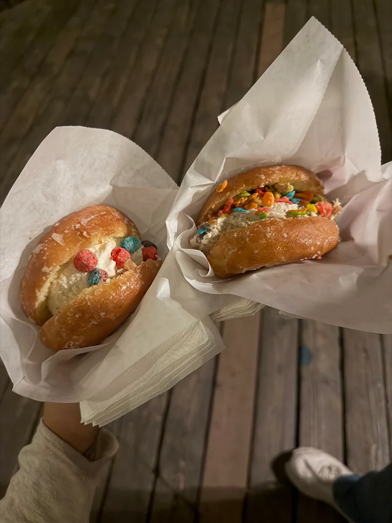 Two hands hold paper-wrapped donuts filled with ice cream and sprinkled with colorful cereal pieces, set against a wooden floor background.