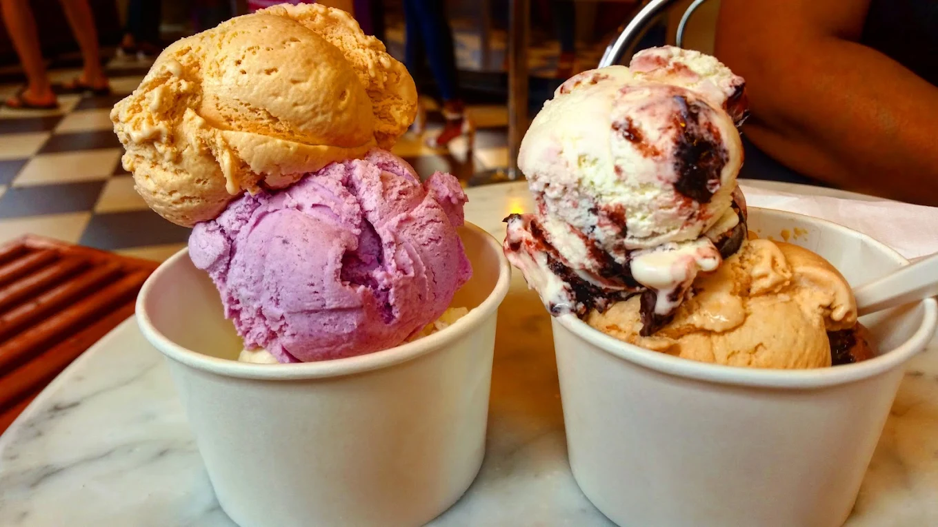 Two cups of colorful ice cream sit on a marble table, each cup filled with three generous scoops in flavors like purple, tan, and swirled white with dark streaks. The background shows a checkered floor and blurred people.