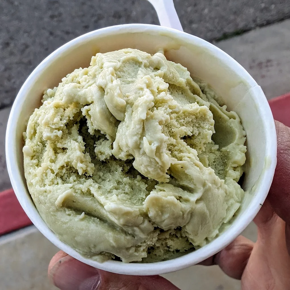 A hand holds a white cup filled with a scoop of light green ice cream. The background shows a sidewalk and a red-painted curb.