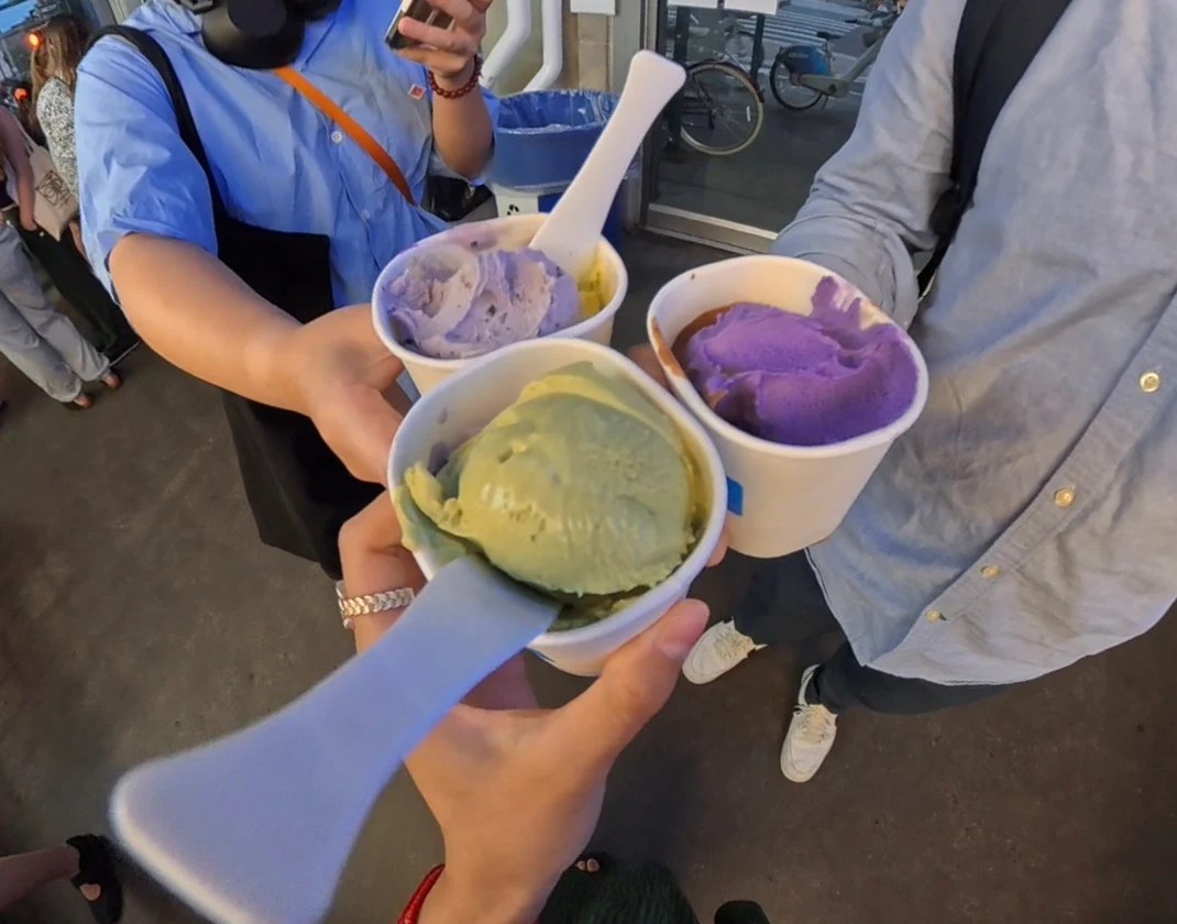 Three people hold ice cream cups with colorful scoops, including green and purple flavors, and white spoons. They are standing outside on a paved area, dressed in casual clothes.