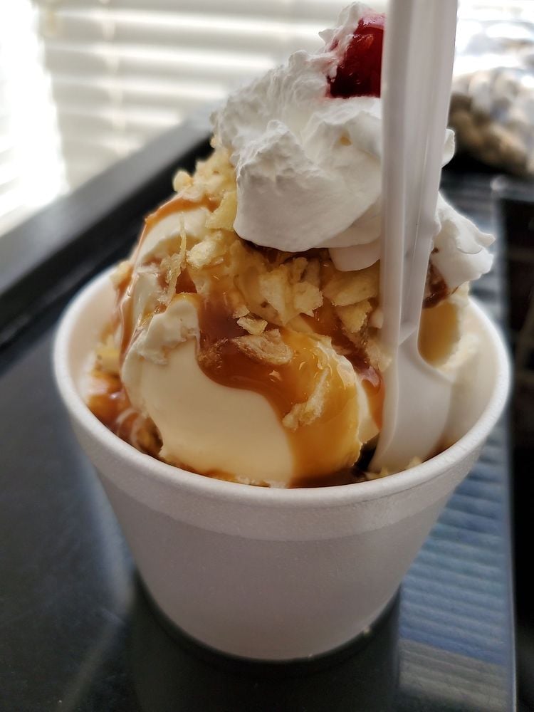 A close-up of a sundae in a white foam cup, topped with whipped cream, caramel sauce, crushed potato chips, and a cherry, with a white plastic spoon inserted. The background shows a window with blinds.
