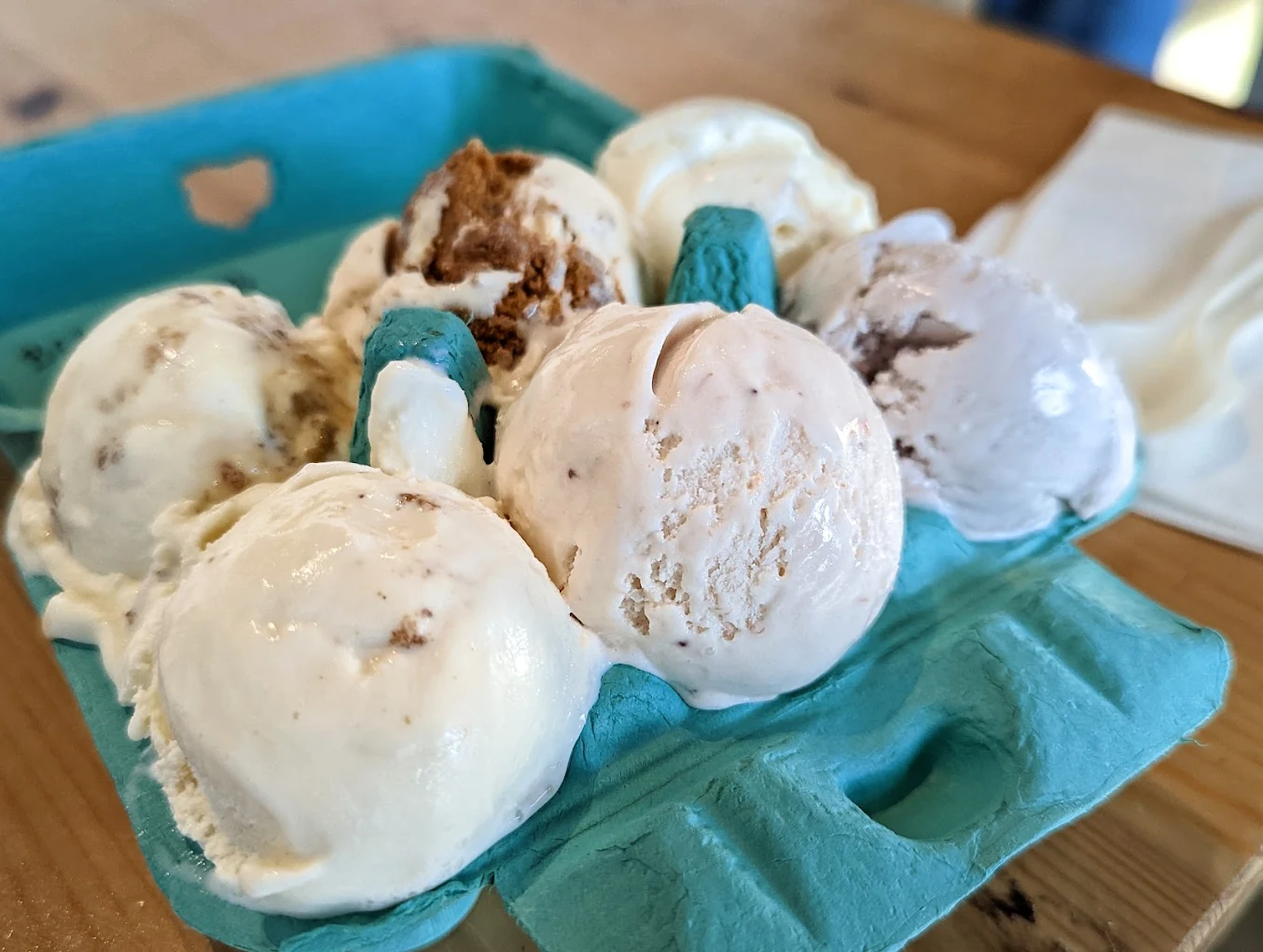 Six scoops of various flavors of ice cream are served in a blue cardboard container, placed on a wooden table with a napkin in the background.
