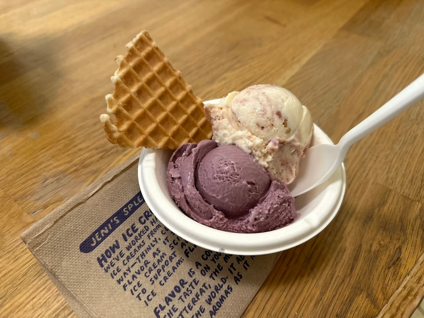 A small paper bowl with two scoops of ice cream—one purple, one white with mix-ins—a waffle cone piece, and a white spoon, sits on a wooden table. A napkin with text is partially visible beneath the bowl.